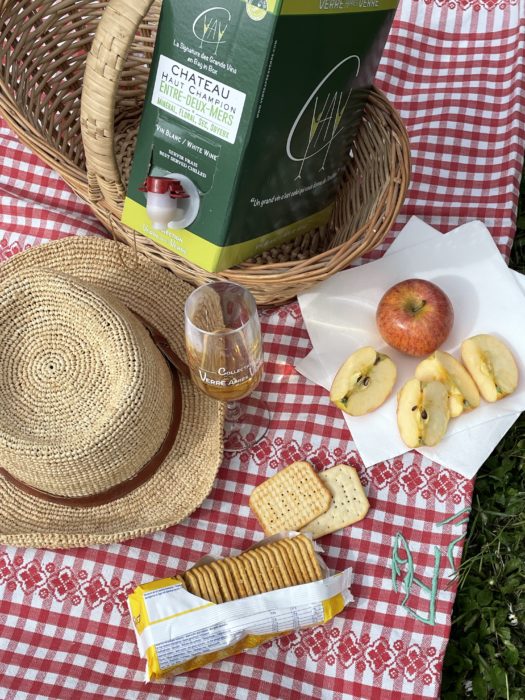 pique-nique sur nappe, panier en osier, chapeau de paille, quartier de pommes, gâteaux apéro et un bag-in-box de vin blanc