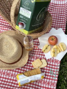Pique-nique sur nappe, panier en osier, chapeau de paille, quartier de pommes, gâteaux apéro et un bag-in-box de vin blanc.